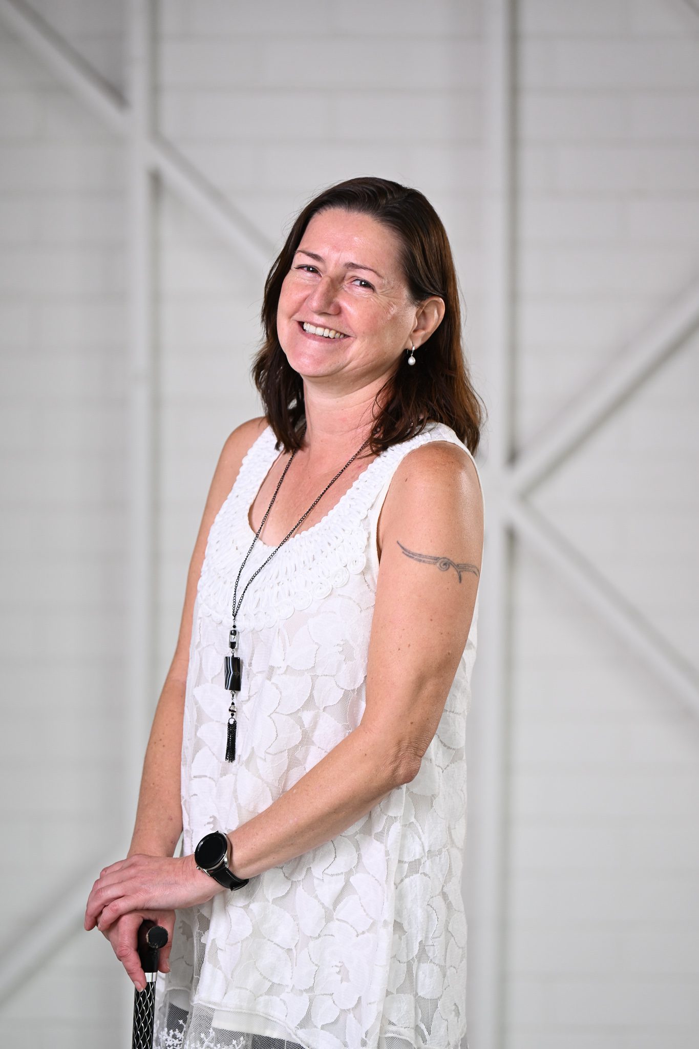 A woman with long brown hair, wearing a white sleeveless blouse, looking at the camera smiling.