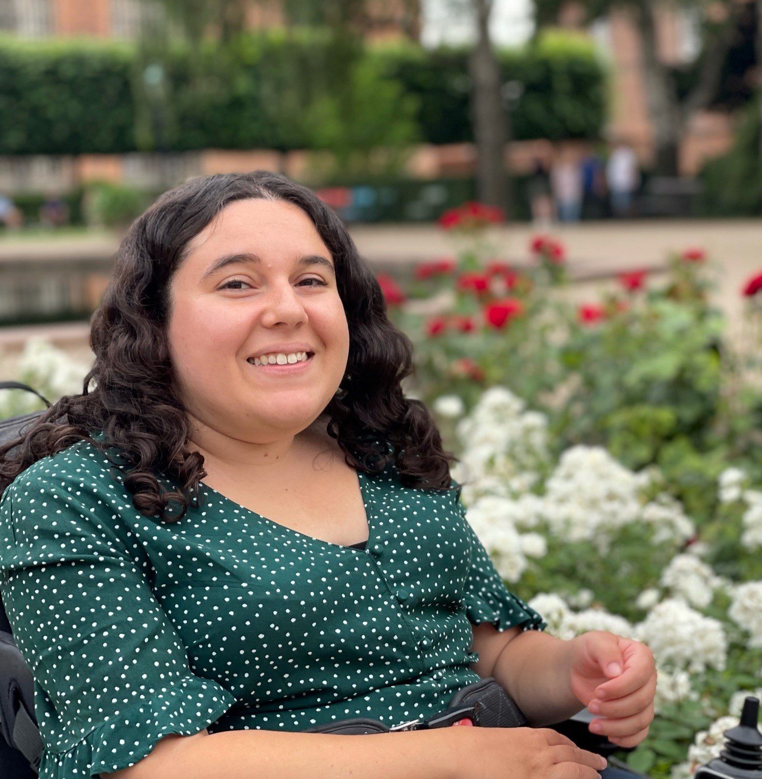 A woman with long brown hair, wearing a green dress and sitting next to a flower garden smiling at the camera.