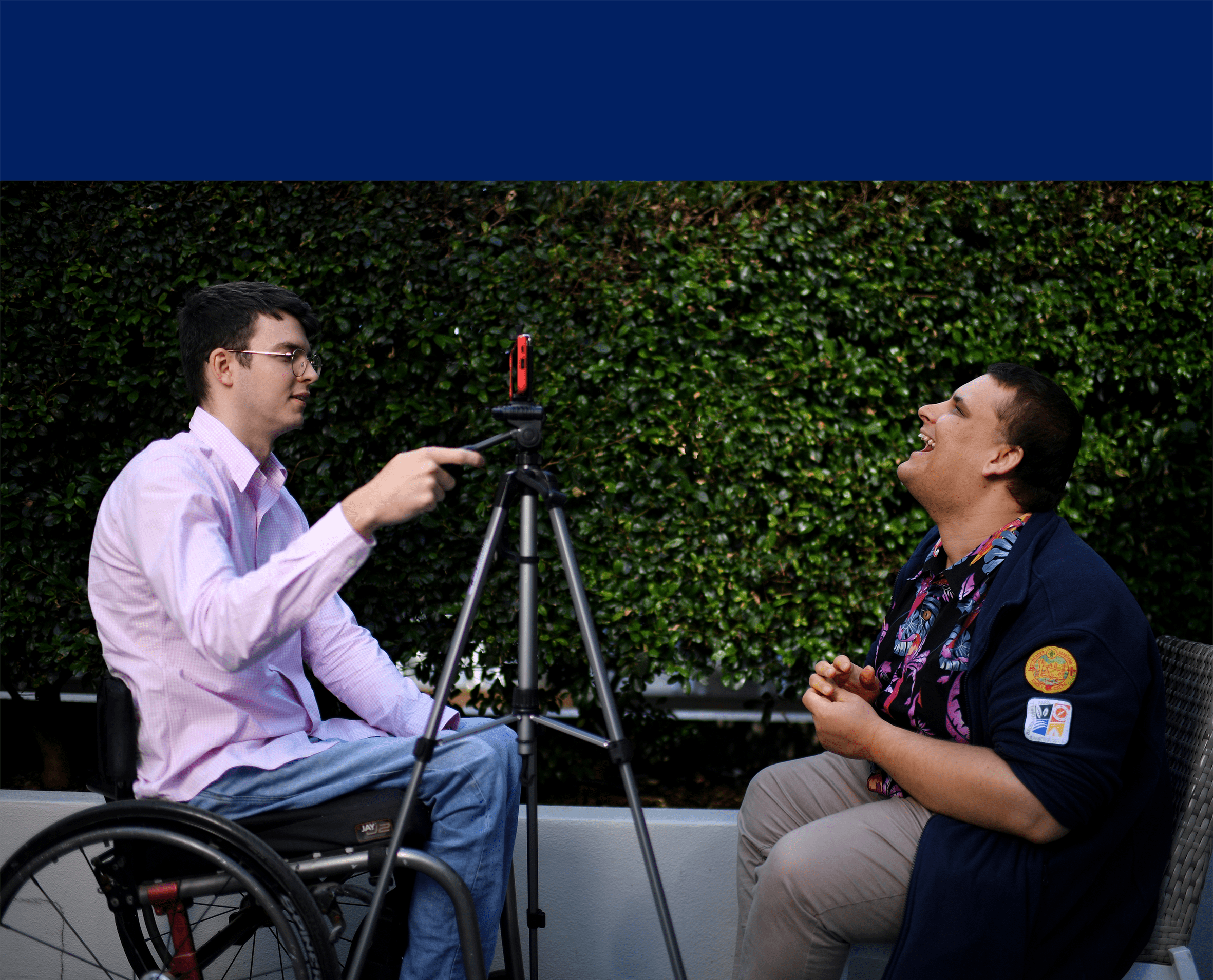 Two men sitting facing each other. One has a mobile on a tripod and recording the other. The man being recorded is laughing.