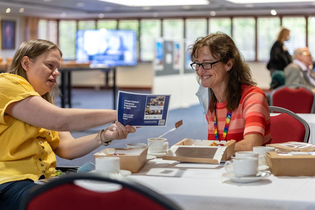 Two women sitting at a table, the one on the left is showing the other a flyer resource and is pointing at it explaining something.