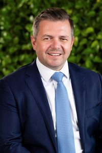 A man with short hair, wearing a navy blue business suit.