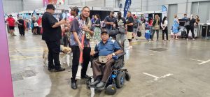 A photo of two people at the Brisbane Disability Connection. One person uses a wheelchair, the other person is standing, holding an Aboriginal musical instrument in is hands. Both people are looking at the camera smiling. 