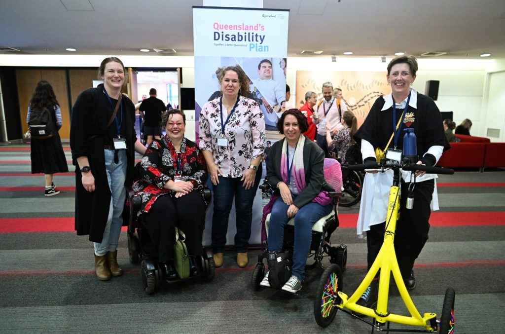 A group of people inside a large grand room. Some people are sitting in wheelchairs, some are standing, and one person is using a bright yellow, stand up mobility device. They are in front of a pull up marketing banner promoting Queensland's Disability Plan. They are looking at the camera smiling.