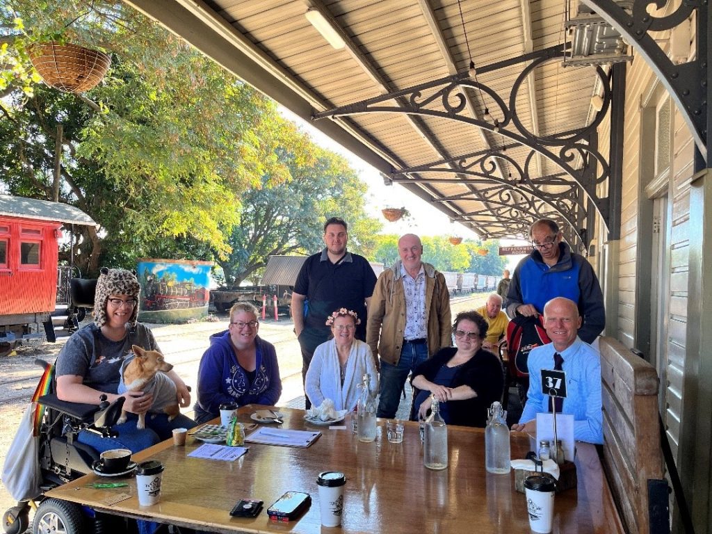 A group of 8 people and a dog sitting around a table outdoors. They appear to be on a veranda, and there is a red train carriage visible in the background. They are all smiling looking at the camera.
